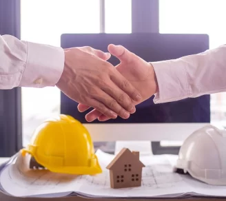 Two people shaking hands with construction blueprints, a notebook, a hard hat, and a small wooden house behind them