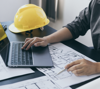 Person using a laptop with blueprints and a hard hat on the table