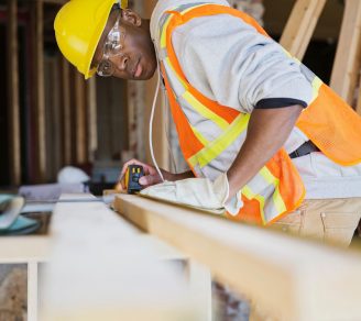 A tradesman in a yellow hard hat measures lumber during construction
